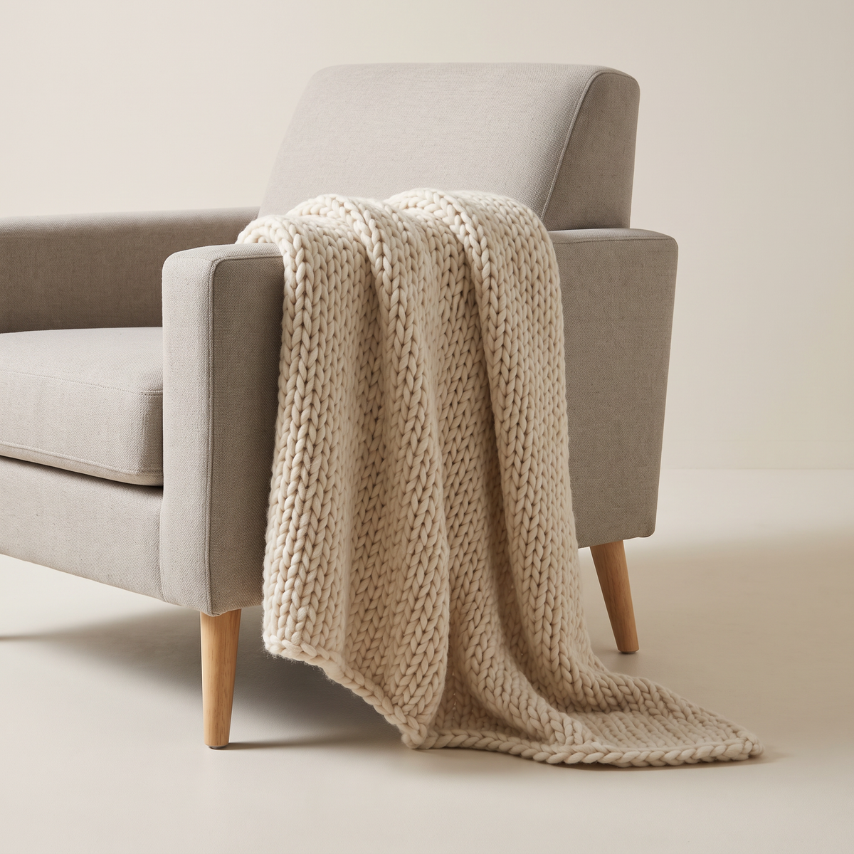Chunky Knit Throw in ivory wool-blend chunky stockinette knit, draped over an armchair arm, front three-quarter view