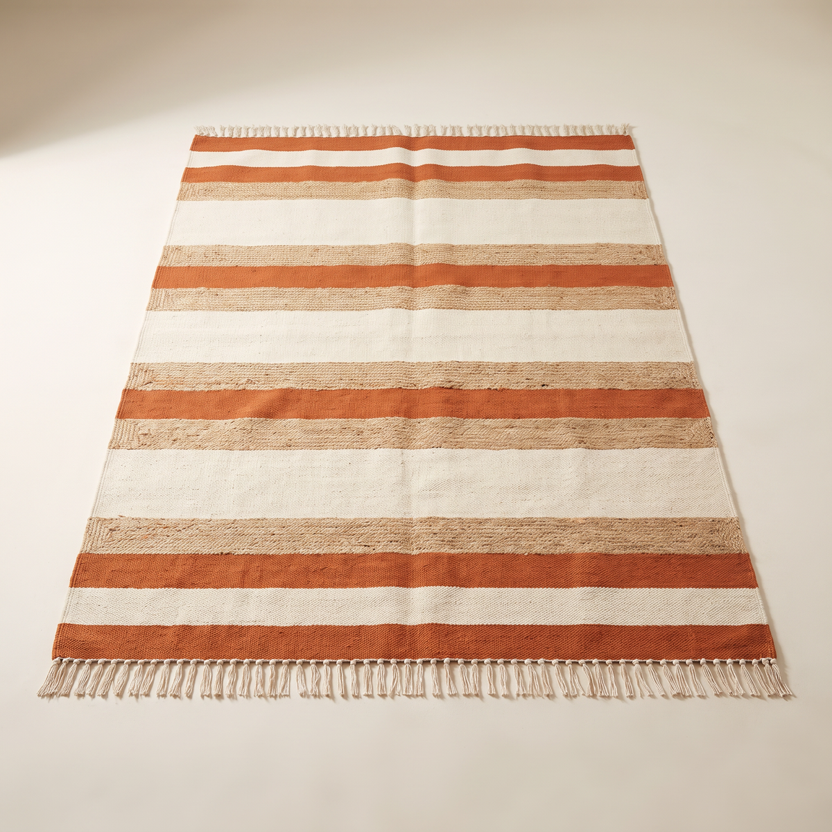Ganga Stripe Dhurrie, flatwoven cotton and jute in ivory, oat and rust bands with knotted fringes, elevated three-quarter view