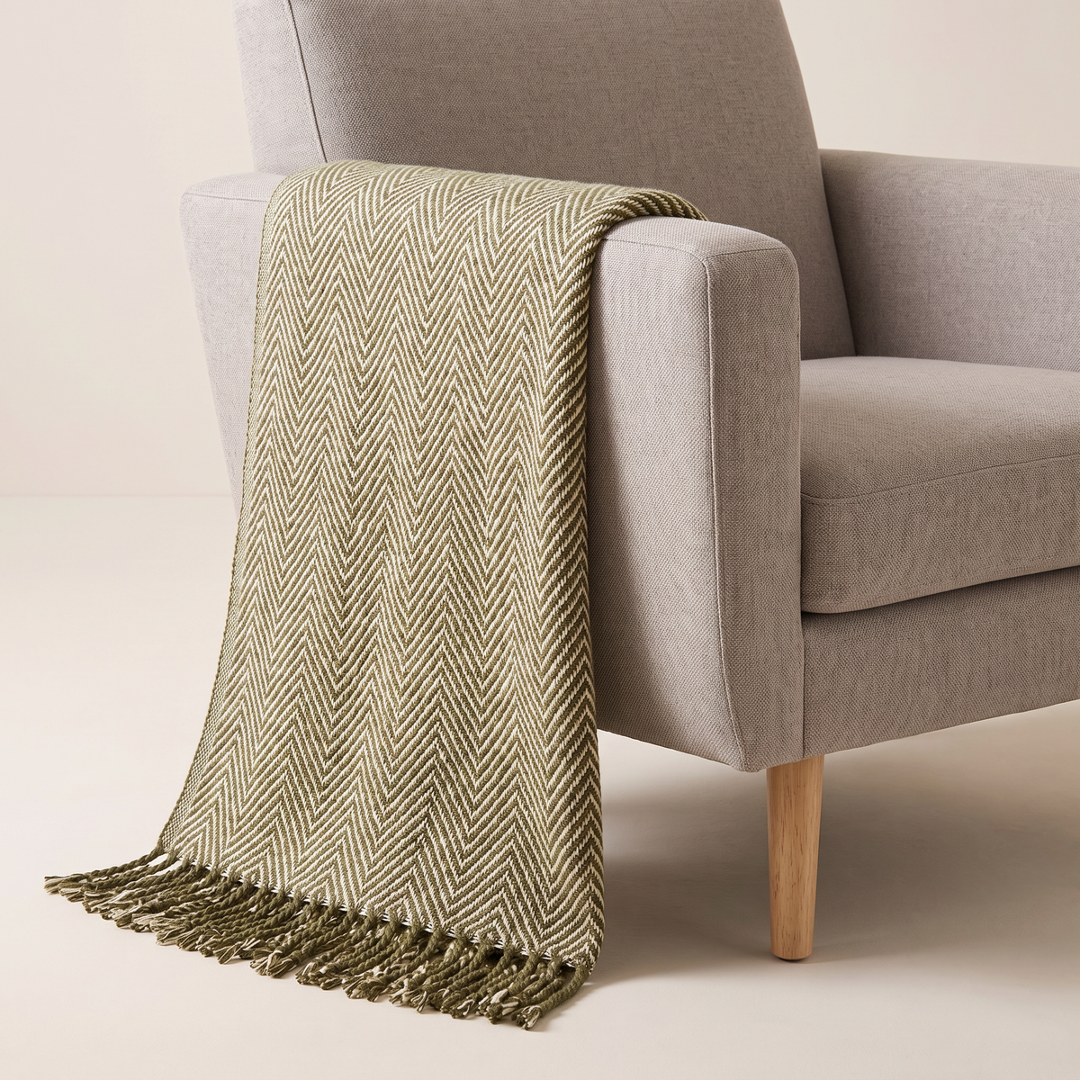 Herringbone Fringe Throw in olive and cream woven herringbone with twisted fringe, draped over an armchair arm, front three-quarter view