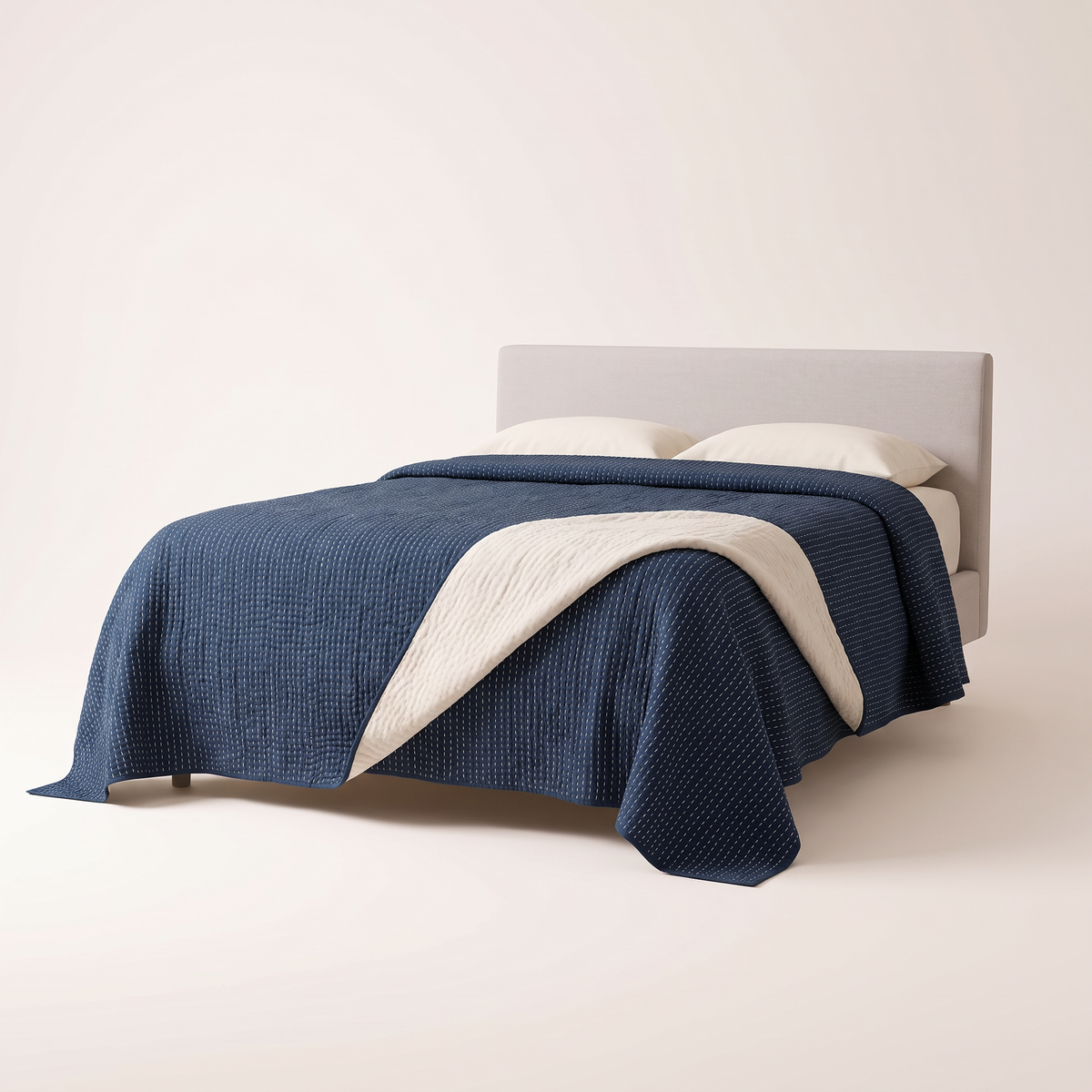 Kantha Quilted Coverlet in deep indigo cotton with rows of ivory running stitch, folded back to show the plain ivory reverse, on a low studio bed, front three-quarter view