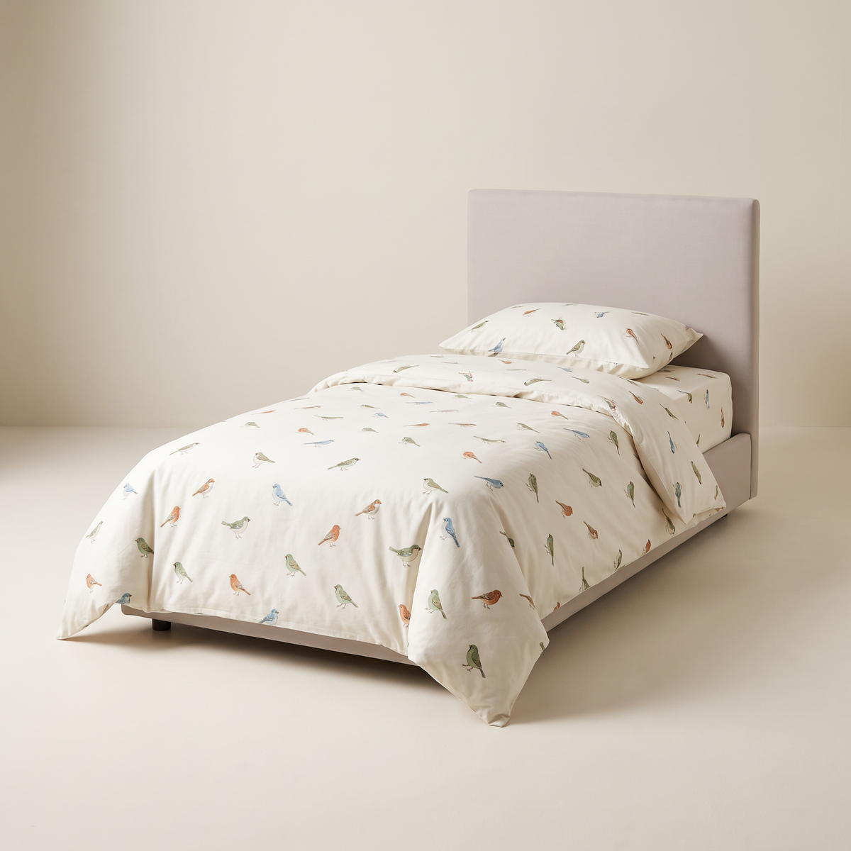 Little Flock Kids Set ivory cotton bedding printed with small sage, rust and sky-blue birds, on a low single studio bed, front three-quarter view