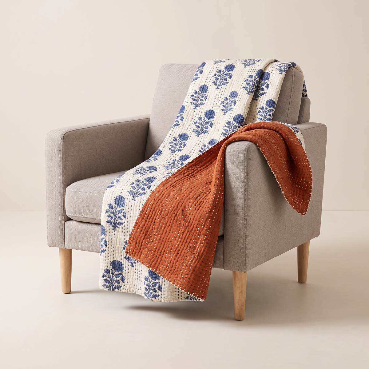 Reversible Kantha Throw with indigo floral face and solid rust reverse in kantha-stitched cotton, draped over an armchair, front three-quarter view
