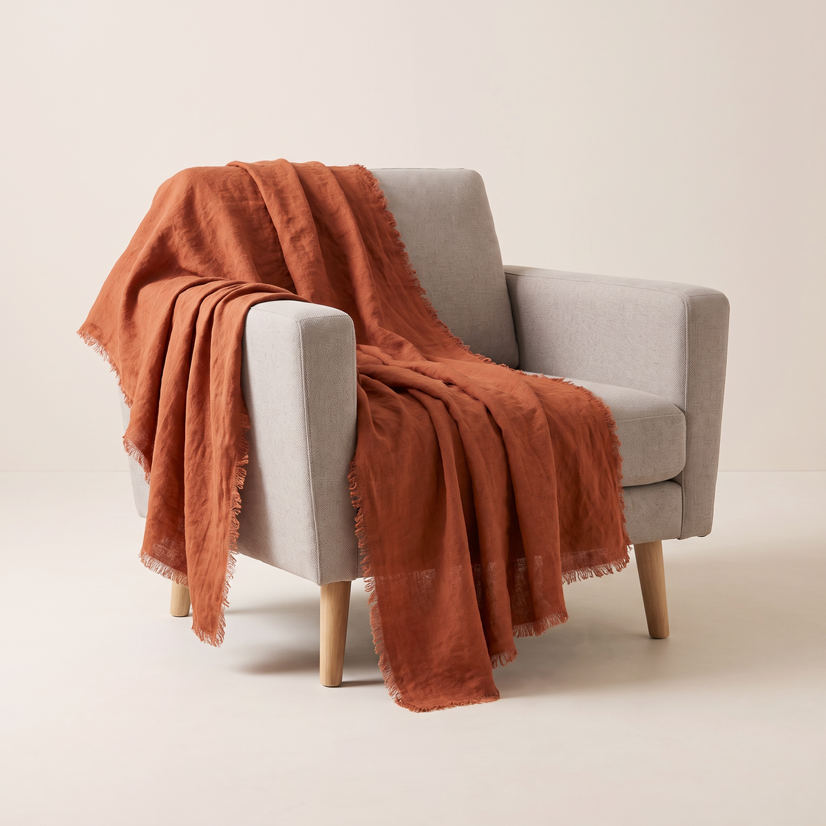 Stonewashed Linen Throw in rust washed linen with frayed edges, draped over an armchair, front three-quarter view