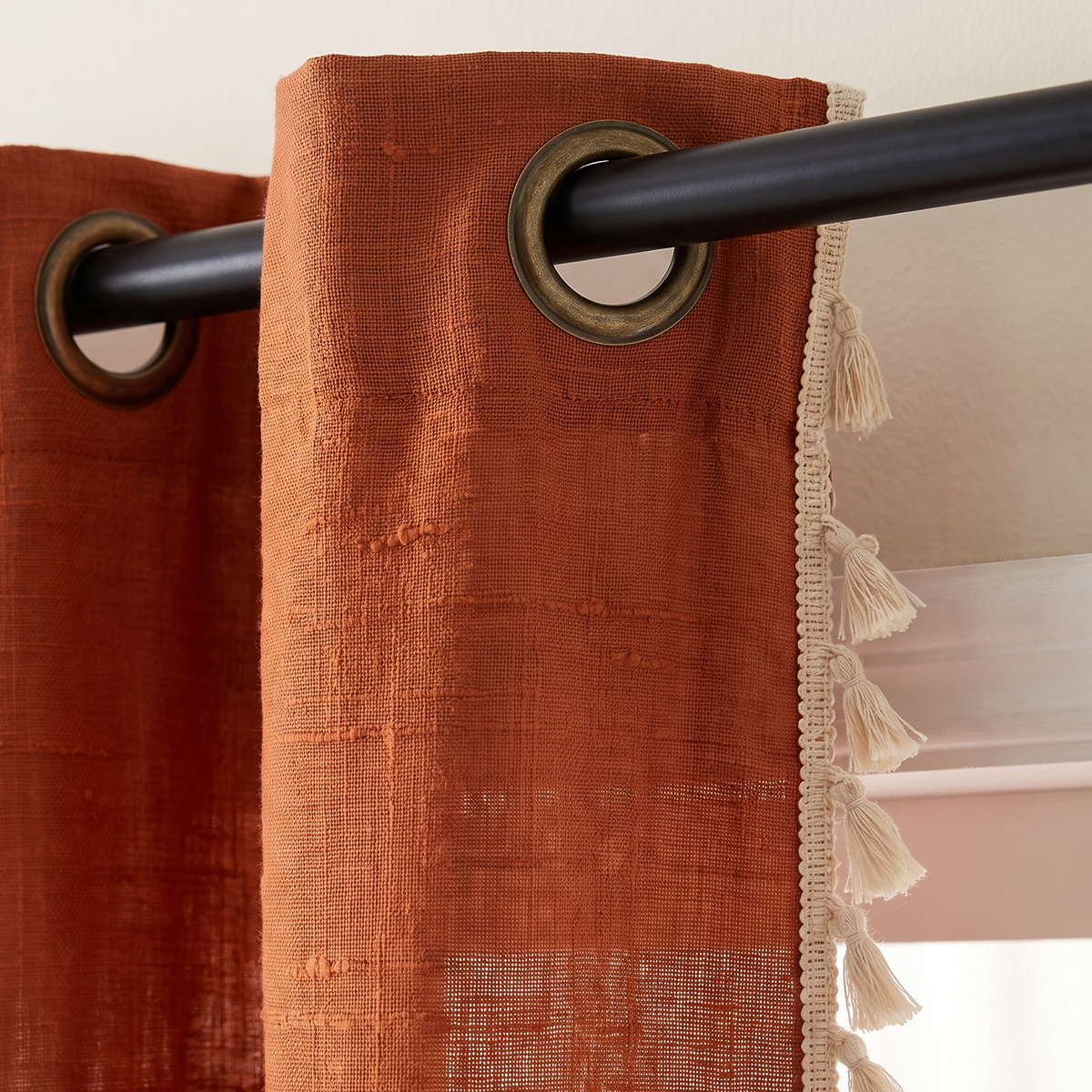 Tassel Slub Eyelet close-up of rust cotton slub weave, antique-brass eyelets on a black rod and hand-tied cream tassel trim