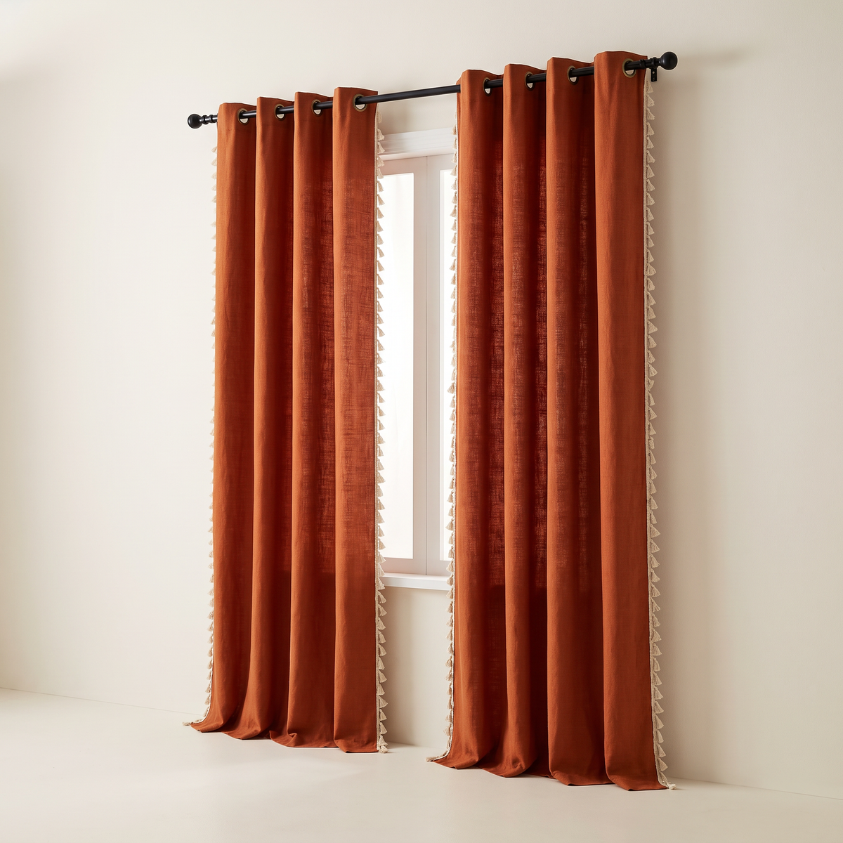 Tassel Slub Eyelet curtains in rust cotton slub with brass eyelets on a black rod and cream tassel trim down both side edges of each panel, front three-quarter view