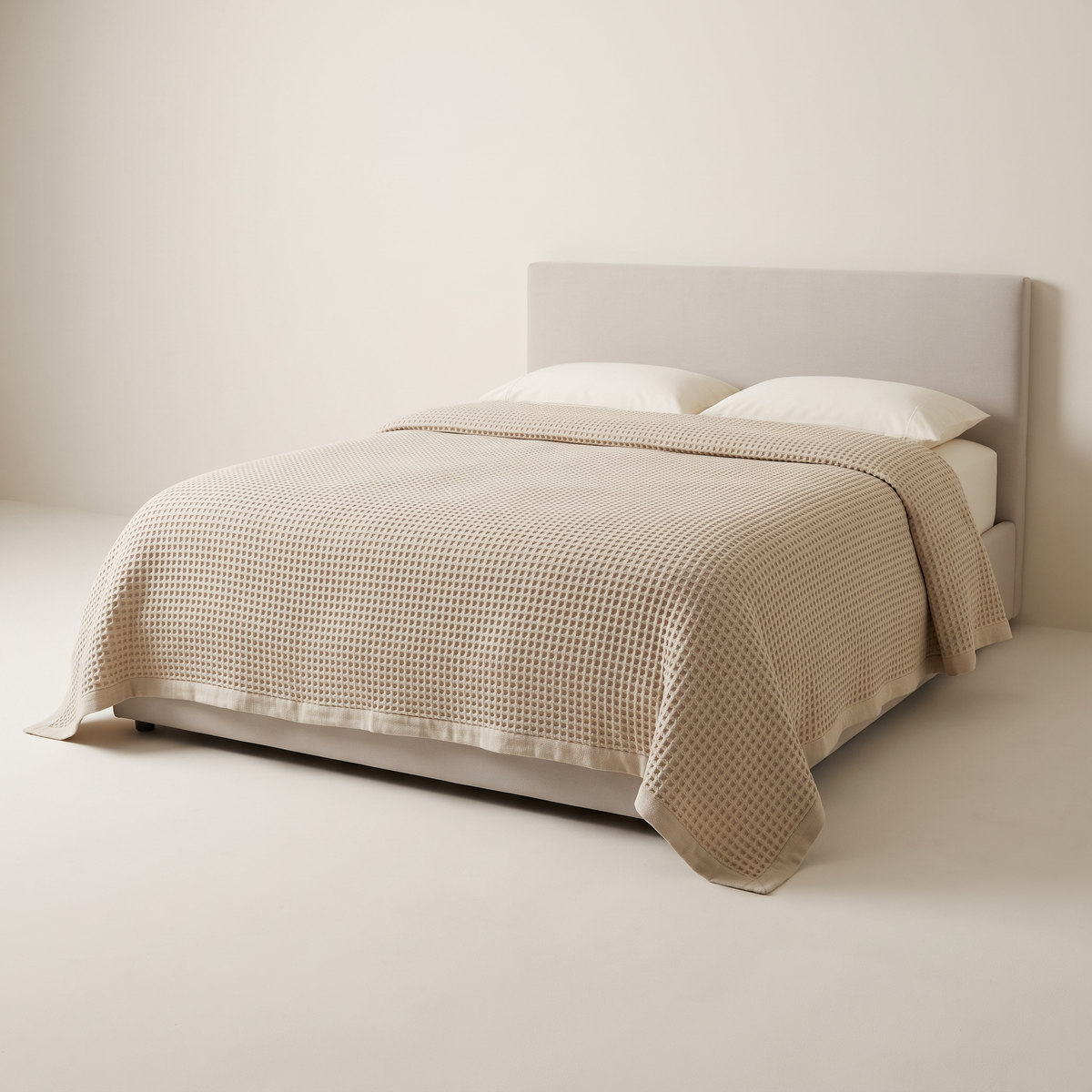 Waffle Weave Bedcover in oat-beige honeycomb waffle cotton with a hemmed edge, on a low studio bed, front three-quarter view